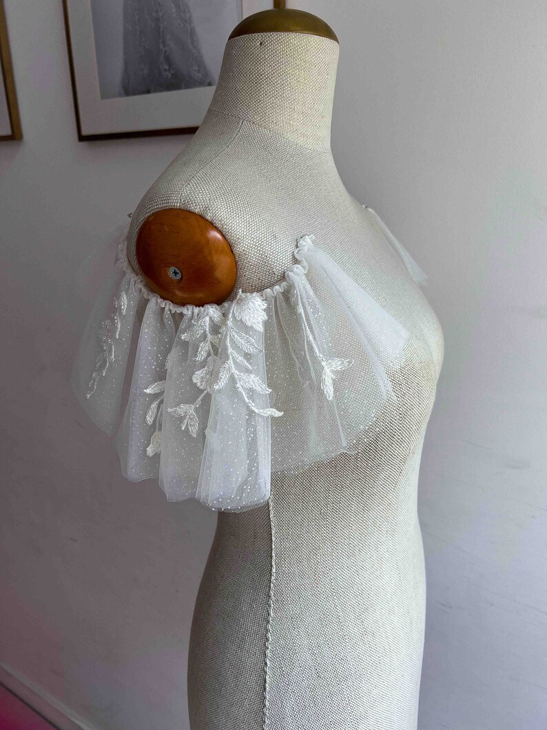 May include: White tulle bridal shoulder piece with delicate floral embroidery and a scalloped edge.