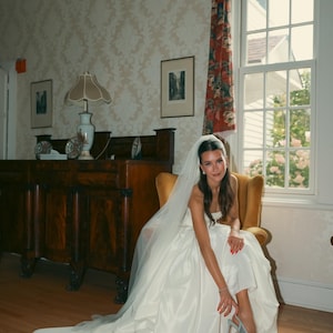K&ouml;nnte beinhalten: Eine Braut in einem wei&szlig;en Satin-Hochzeitskleid mit langer Schleppe sitzt in einem gelben Stuhl. Sie tr&auml;gt einen Schleier und silberne Abs&auml;tze. Der Raum hat ein gro&szlig;es Fenster mit floralen Vorh&auml;ngen und antiken M&ouml;beln.