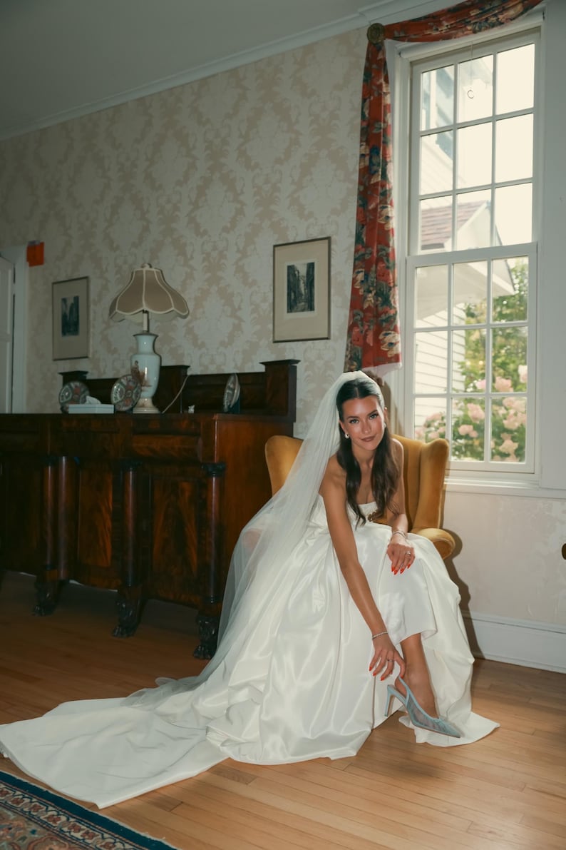 K&ouml;nnte beinhalten: Eine Braut in einem wei&szlig;en Satin-Hochzeitskleid mit langer Schleppe sitzt in einem gelben Stuhl. Sie tr&auml;gt einen Schleier und silberne Abs&auml;tze. Der Raum hat ein gro&szlig;es Fenster mit floralen Vorh&auml;ngen und antiken M&ouml;beln.