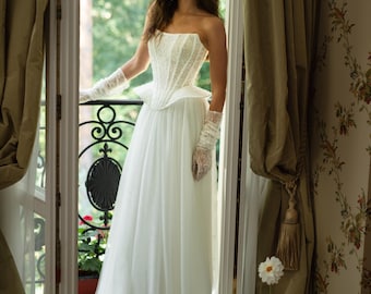 Peut inclure: Robe de mariée blanche sans bretelles avec un corsage perlé et une jupe fluide. La robe est assortie de gants en dentelle. La scène se déroule dans une porte avec des rideaux floraux et une vue sur la verdure.
