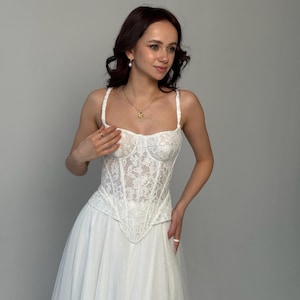 May include: A white lace corset top featuring a sweetheart neckline and thin shoulder straps, paired with a long, flowing white skirt. The corset bodice has a pointed hemline and intricate floral lace detailing throughout the fabric.
