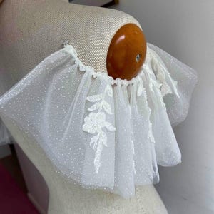 May include: White tulle fabric with a delicate floral pattern and a ruffled edge. The fabric is sheer and has a dotted texture.