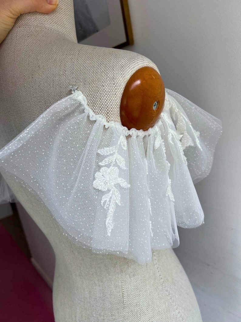 May include: White tulle fabric with a delicate floral pattern and a ruffled edge. The fabric is sheer and has a dotted texture.