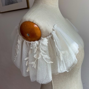 May include: White tulle bridal shoulder piece with delicate floral embroidery and a subtle sparkle.