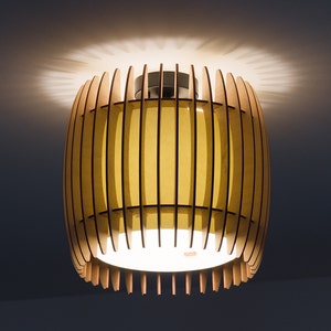 Può includere: Un apparecchio di illuminazione a soffitto rotondo in legno con una finitura marrone chiaro e una luce bianca che brilla attraverso le lamelle.