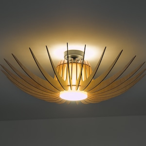 Może przedstawiać: Nowoczesna lampa sufitowa wykonana z drewna w jasnobrązowym wykończeniu. Lampa ma okrągły kształt z wieloma drewnianymi listwami promieniującymi na zewnątrz, tworząc wzór gwiazdy. Lampa jest oświetlona ciepłym białym światłem.