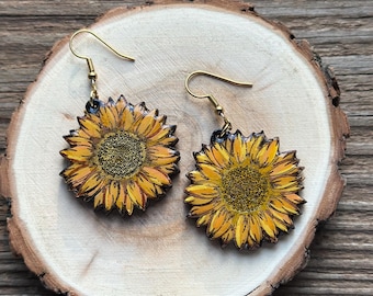 Könnte beinhalten: Ein Paar Sonnenblumenohrringe mit goldfarbenen Haken. Die Ohrringe sind mit gelben und braunen Details bemalt, die das Aussehen einer Sonnenblume nachahmen. Die Ohrringe werden auf einer Holzscheibe präsentiert.