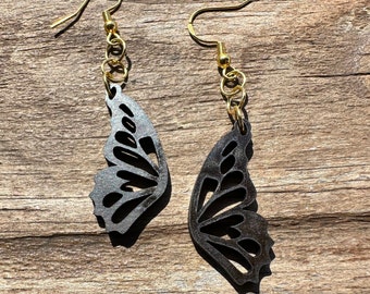 Puede incluir: Un par de pendientes negros en forma de mariposa con ganchos y cadenas doradas. Los pendientes están hechos de madera y tienen un diseño delicado e intrincado.