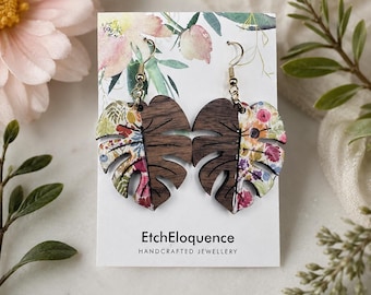 May include: A pair of monstera leaf-shaped earrings featuring a split design with one half in dark brown wood grain and the other half in a colorful floral pattern. The earrings hang from gold-toned metal hooks on a white display card with text.
