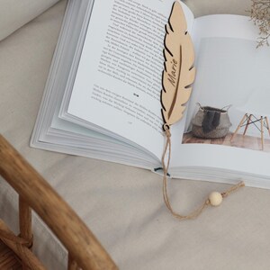 Könnte beinhalten: Ein Holz-Feder-Lesezeichen mit dem eingravierten Namen "Marie". Das Lesezeichen ist an einer Schnur mit einer Holzperle befestigt. Das Lesezeichen liegt auf einem geöffneten Buch.