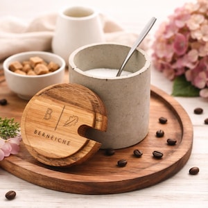 Könnte beinhalten: Ein Holzserviertablett mit einer Zuckerdose, Zuckerwürfeln und einem Betonzuckerbehälter mit einem Löffel. Ein Holzdeckel mit dem Text "BERNEICHE" bedeckt die Zuckerdose. Kaffeebohnen und rosa Blumen sind verstreut.