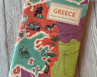 May include: Vintage travel guide titled "Greece" from the American Geographical Society's "Around the World Program." The cover features a colorful map of Greece with illustrations of people and landmarks. The book is set against a light wood grain background.