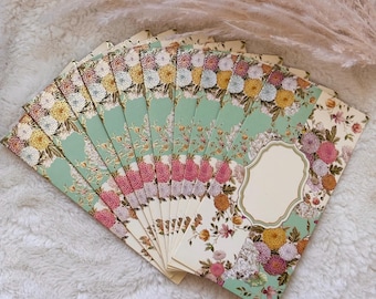 Puede incluir: Un abanico de sobres rectangulares de papel con un estampado floral de crisantemos amarillos, rosas y blancos sobre un fondo verde claro y crema. Cada sobre tiene una etiqueta en blanco con bordes festoneados para escribir.