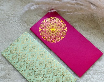Puede incluir: Dos sobres decorativos. Uno es rosa fucsia con un diseño de mandala dorado. El otro es verde claro con un patrón de cachemira dorado. Ambos son rectangulares y están sobre una superficie blanca y esponjosa.
