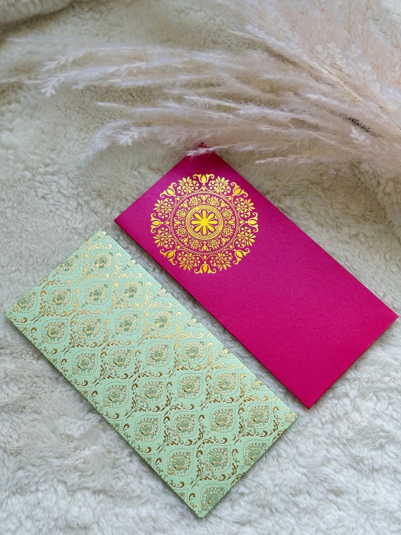 May include: Two decorative envelopes with gold and green patterns. One envelope is pink with a gold mandala design, and the other is light green with a gold paisley pattern.