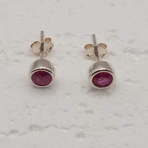 May include: A pair of stud earrings featuring round, deep pink gemstones set in polished silver-toned metal. Each earring has a smooth, circular bezel setting and includes a matching silver-toned post back with a small butterfly clutch.