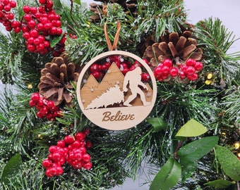 May include: A wooden Christmas ornament featuring a silhouette of Bigfoot walking through a snowy forest with the word "Believe" inscribed below. The ornament is hanging on a green Christmas tree branch with red berries.