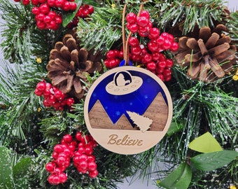 May include: A wooden Christmas ornament with a blue background and a silhouette of a UFO flying over mountains. The ornament has the word "Believe" written on the bottom.