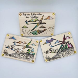 May include: Three rectangular wooden plaques with a laser-etched design. Each features a glider soaring above a landscape with mountains, clouds, and the text "Auf ein Leben über den Wolken!". Gold-colored rivets secure the corners.