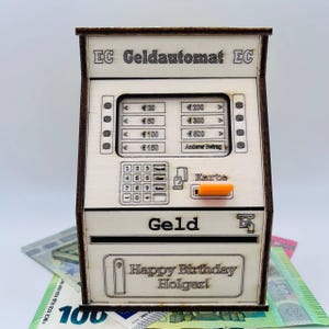 Könnte beinhalten: Eine hölzerne Box in Form eines Geldautomaten mit dem Text "EC Geldautomat EC" oben. Die Vorderseite hat Knöpfe, einen Kartensteckplatz und ein Display mit Euro-Beträgen. Der Boden trägt den Text "Happy Birthday Holger!" und das Wort "Geld".