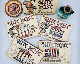 May include: Five wooden plaques with gold accents, each featuring the text "Gute Reise" in a stylised font, with the words "nach Griechenland" below. Each plaque has a different image cut out of the wood, including a map of Greece, a Greek temple, and a tree.