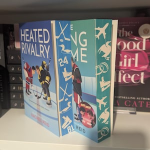 Könnte beinhalten: Zwei Bücher mit den Titeln "Heated Rivalry" und "The Long Game" werden auf einem Regal ausgestellt. Das Buch "Heated Rivalry" zeigt Eishockeyspieler auf dem Cover. Das andere Buch hat einen Eishockeyspieler auf dem Cover. Die Bücher stehen auf einem weißen Regal mit anderen Büchern.