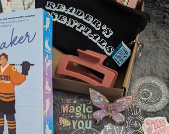 May include: A box filled with various items for book lovers, including a book titled "The Baker", a black zippered pouch with the text "Reader's Essentials", a pink hair clip, and several holographic stickers with book-themed designs. The stickers feature phrases like "The Magic is in You", "Read All The Books", and "Sorry, I'm Booked".