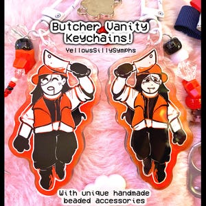 May include: Two acrylic keychains featuring a cartoon butcher character with a cleaver, wearing an orange jacket and hat. The keychains have unique handmade beaded accessories and the text "Butcher Vanity Keychains!" and "YellowsSillySymphs".