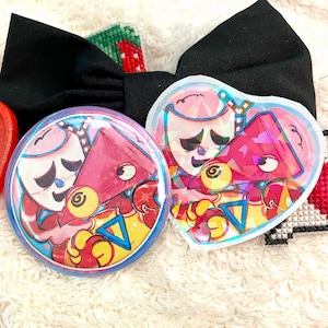 Pode incluir: Dois pins holográficos com uma personagem de desenho animado com cabeça triangular rosa e um triângulo azul no peito, um circular e outro em forma de coração, posicionados à frente de um grande laço de tecido preto e outros pequenos artigos de ponto cruz.
