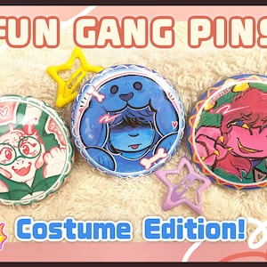 Peut inclure: Trois badges ronds illustrés de personnages colorés. Le badge de gauche montre une créature blanche à cornes en tenue verte. Le badge central représente un personnage bleu à capuche. Le badge de droite présente un personnage rose.