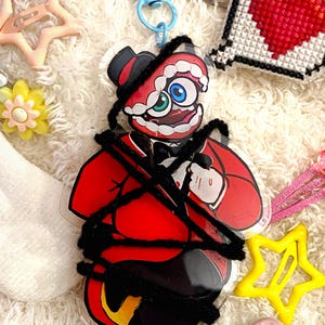 Puede incluir: Un dije acrílico transparente con un personaje de dibujos animados rojo y negro con sombrero de copa y pajarita, sujeto a un cierre azul. El dije está rodeado de accesorios para el cabello, incluidos clips en forma de estrella y flor. Otros artículos incluyen un corazón bordado.