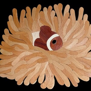 Könnte beinhalten: Ein Wandbild aus Holz, das einen Clownfisch in einer Seeanemone zeigt. Der Fisch ist aus dunkelrotbraunem und hellem cremefarbenem Holz gefertigt, während die Anemone aus zahlreichen geschichteten, hellbraunen Holzstreifen besteht.