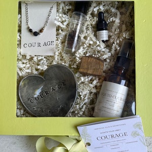 May include: A yellow gift box filled with self-care items. Includes a necklace, heart-shaped dish with "COURAGE" text, essential oil bottles, and a card. The box contains shredded paper for cushioning.