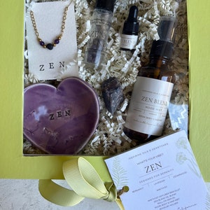 May include: A yellow gift box with a clear lid, filled with a variety of items, including a purple heart-shaped dish with the word "ZEN" on it, a small bottle of essential oil, a spray bottle, a necklace, a crystal, and a card that reads "ZEN" and "Designed for Serenity".