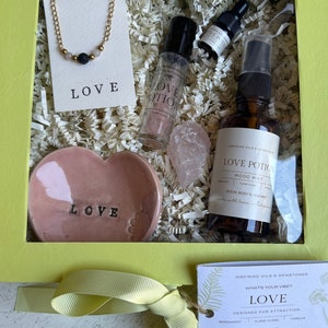 May include: A gift box with a yellow frame contains a gold chain necklace with beads, a pink heart-shaped dish with "LOVE" text, and several bottles labeled "LOVE POTION". The box includes a tag that says "LOVE" and "Designed for Attraction".