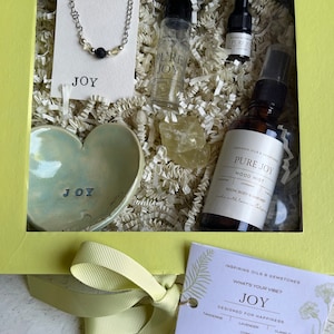 May include: A yellow gift box with a green interior contains a variety of items, including a citrine crystal, a diffuser bracelet, a roll-on essential oil blend, a room and body mist, and a jewelry holder. The box is tied with a yellow ribbon and has a tag that reads "Inspiring Oils & Gemstones What's Your Vibe? Joy Designed for Happiness Tangerine Lavender Citrine Ylang Ylang Citrine Diffuser Bracelet Pure Joy Oil Blend Roll-on Gem Oil Room Body & Hair Mist Jewelry Holder Made with Love in California".
