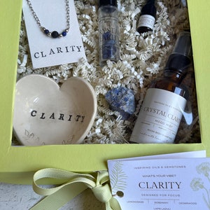 May include: A yellow gift box with a light green ribbon. The box contains a heart-shaped dish, a small bottle of essential oil, a crystal, a necklace, and a card that says "Crystal Clarity Mood Mist". The card also says "Inspiring Oils & Gemstones" and "What's Your Vibe? Clarity".
