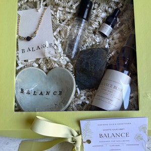 May include: A green gift box with a clear window showing a heart-shaped dish with the word "BALANCE" on it, a black stone, a gold necklace with black beads, and three bottles of essential oil. The box also includes a tag with the words "BALANCE" and "INSPIRING OILS & GEMSTONES".