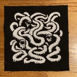 May include: Square black patch featuring a white illustration of intertwined, worm-like creatures with open mouths and sharp teeth. The design is densely packed, creating a textured appearance. The patch is likely for clothing or accessories.