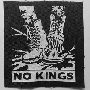 May include: A square black and white fabric patch featuring a graphic illustration of two lace-up boots standing above a crown. The design includes a stylized dripping effect beneath the boots and the text NO KINGS printed in a bold font at the bottom.