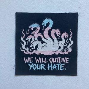 May include: A square black fabric patch featuring an embroidered multi-headed hydra creature in light blue, white, and pink gradients. Below the creature, the text "WE WILL OUTLIVE YOUR HATE." is embroidered in matching light blue and pink colors.