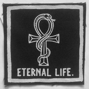May include: A square black fabric patch with a white embroidered border. The center features a white embroidered ankh symbol entwined with a snake. Below the symbol, the text ETERNAL LIFE. is embroidered in white capital letters.