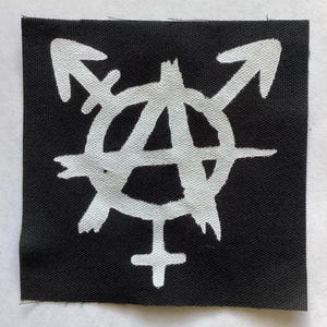 May include: Square black patch with a white anarchist symbol incorporating gender symbols and arrows. The design is hand-painted, with a rough, textured appearance. The patch is likely for clothing or accessories.