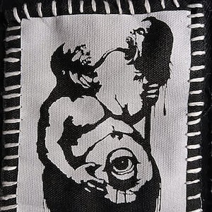 May include: A black and white patch featuring a graphic illustration of two figures interacting, with a large eye at the bottom. The patch is stitched onto a black fabric with white thread.
