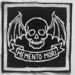 May include: A square fabric patch featuring a black background with a white embroidered design. The graphic depicts a central human skull with bat-like wings extending from the sides. Below the skull is a banner with the text MEMENTO MORI.