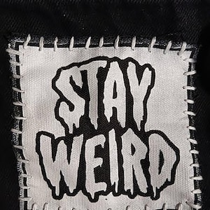 May include: A black fabric patch with the words "STAY WEIRD" in bold, dripping-style white letters. The patch is stitched onto a black background with white thread, creating a contrasting border.