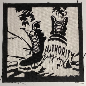 May include: A square black and white fabric patch featuring a high-top boot graphic and the word AUTHORITY printed in a bold, stylized font. The design includes abstract, jagged black ink-like shapes surrounding the boot and text on a white background.