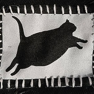 May include: Black cat silhouette patch on a white background, stitched onto black fabric. The cat is in mid-leap, with its tail curved. The patch is framed with white stitching.