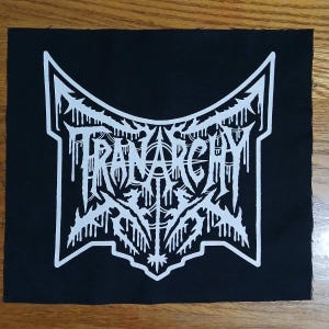 May include: Black square patch with a white graphic design. The design features the word "TRANARCHY" in a stylized, gothic font, surrounded by a symmetrical, ornate border with dripping elements. The patch is likely for clothing or accessories.