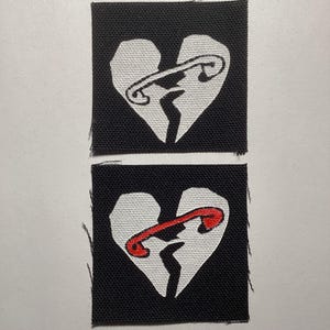 May include: Two square black fabric patches featuring a white broken heart graphic. The top patch shows a white safety pin holding the heart together, while the bottom patch features a red safety pin holding the white broken heart together.